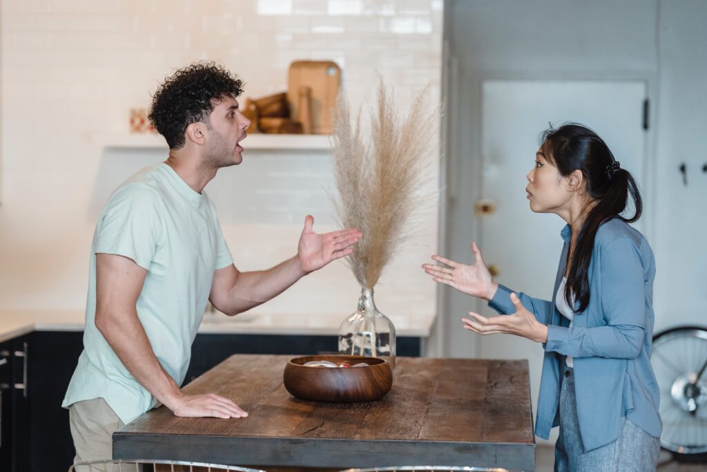 White man and Asian woman standing up arguing across a kitchen counter. They may want to seek therapy for adults in Sheffield.